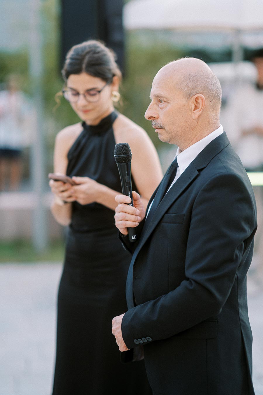 Man in a suit holding a microphone, with a woman in a black dress using a smartphone in the background, during an outdoor event.