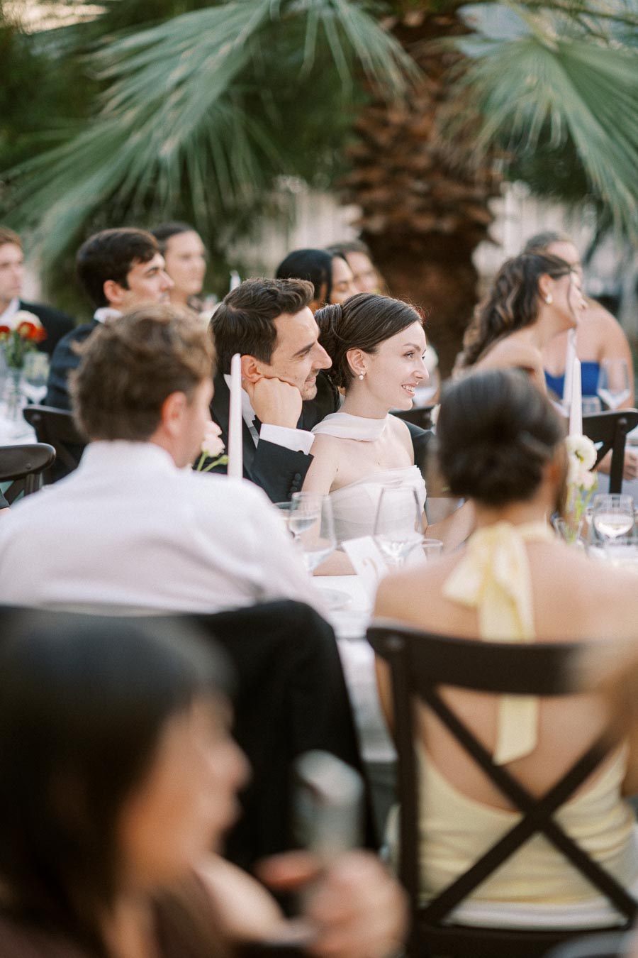 Elegant wedding reception with guests seated at a beautifully decorated outdoor table, featuring lush palm trees in the background and a joyful couple enjoying the celebration.