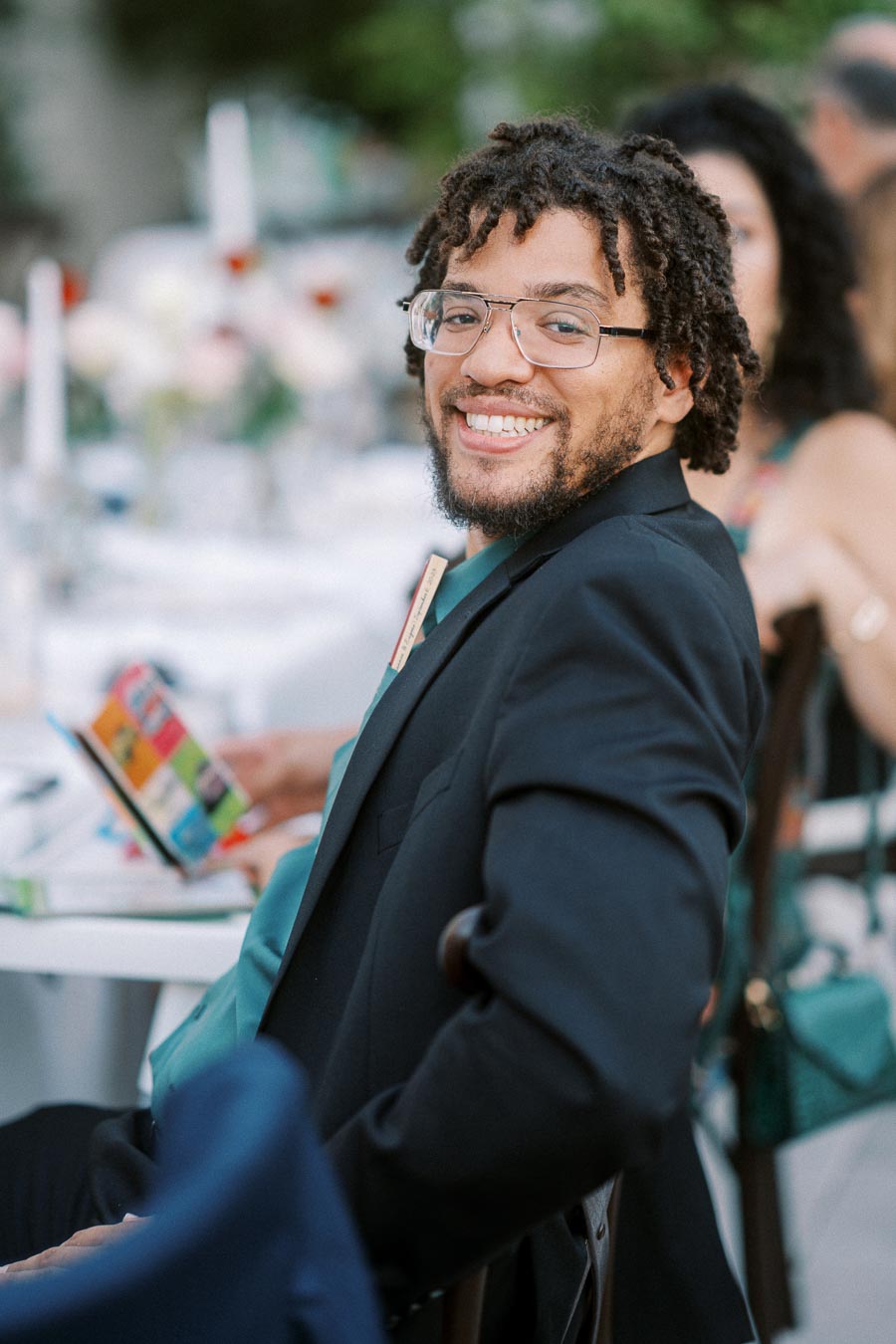 A smiling man with curly hair and glasses wearing a black suit jacket seated at an outdoor event.