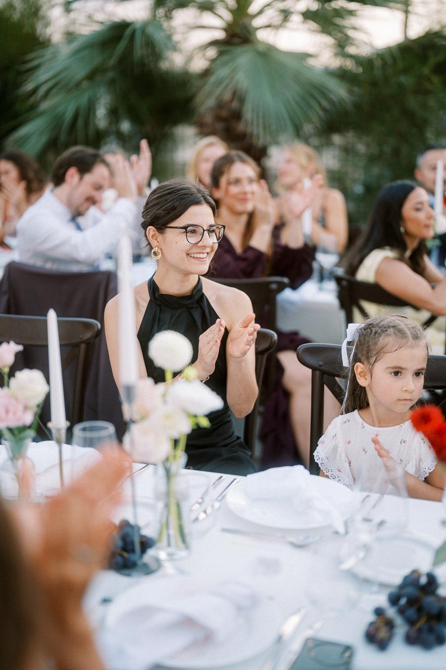 Woman and child clapping at an elegant outdoor event with floral decorations and palm trees in the background.
