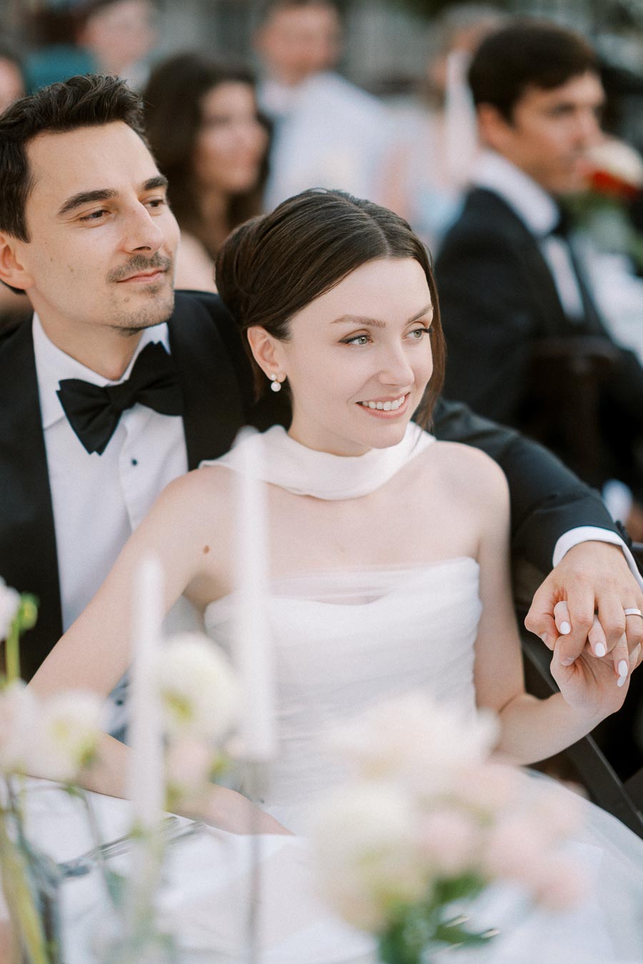 A smiling bride and groom holding hands at their wedding reception, surrounded by soft-focus floral decorations and elegantly dressed guests.