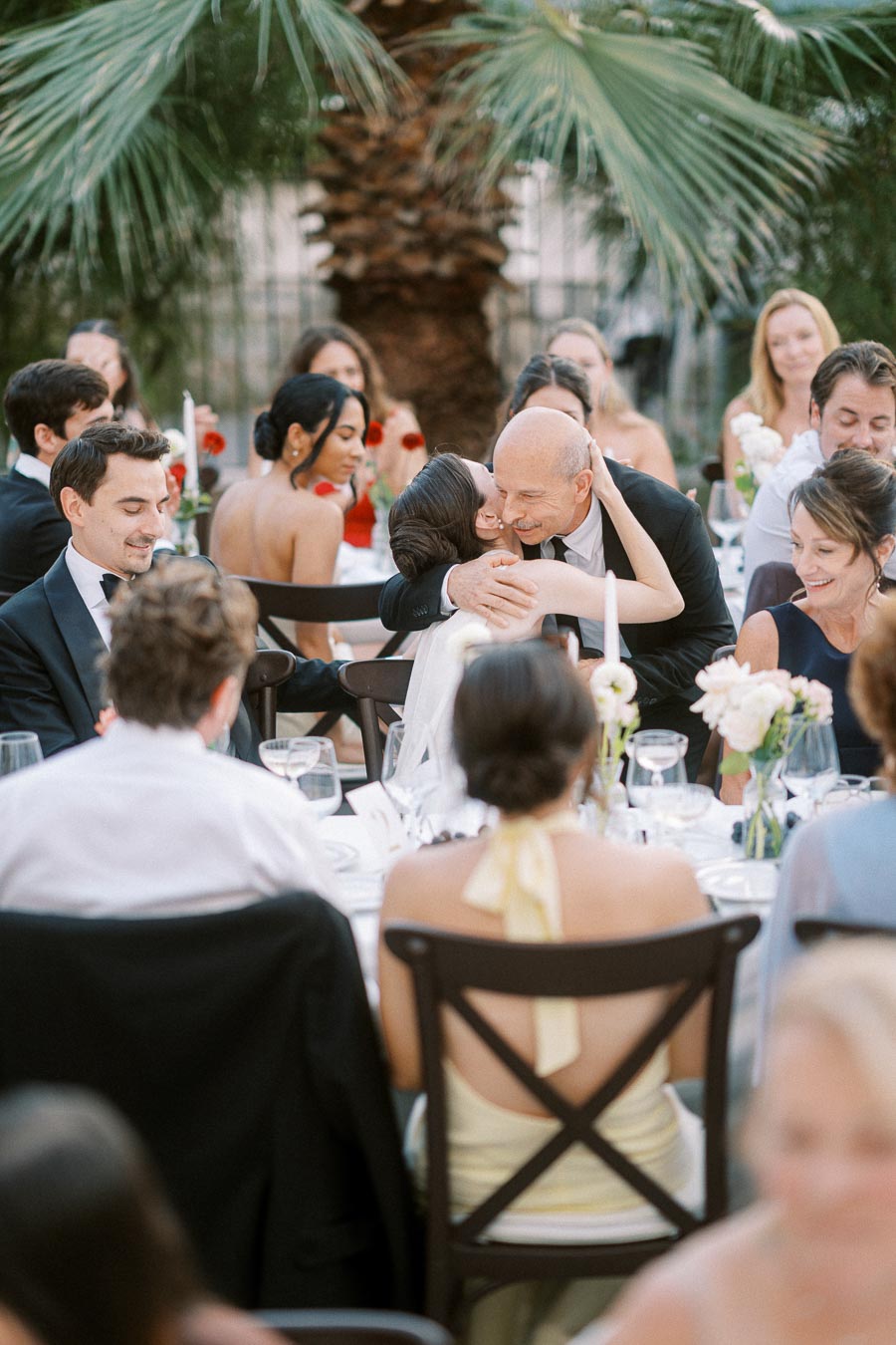 A joyful outdoor wedding reception with guests celebrating; a woman in a dress hugs a man in a suit amid elegantly set tables adorned with flowers and surrounded by palm trees.