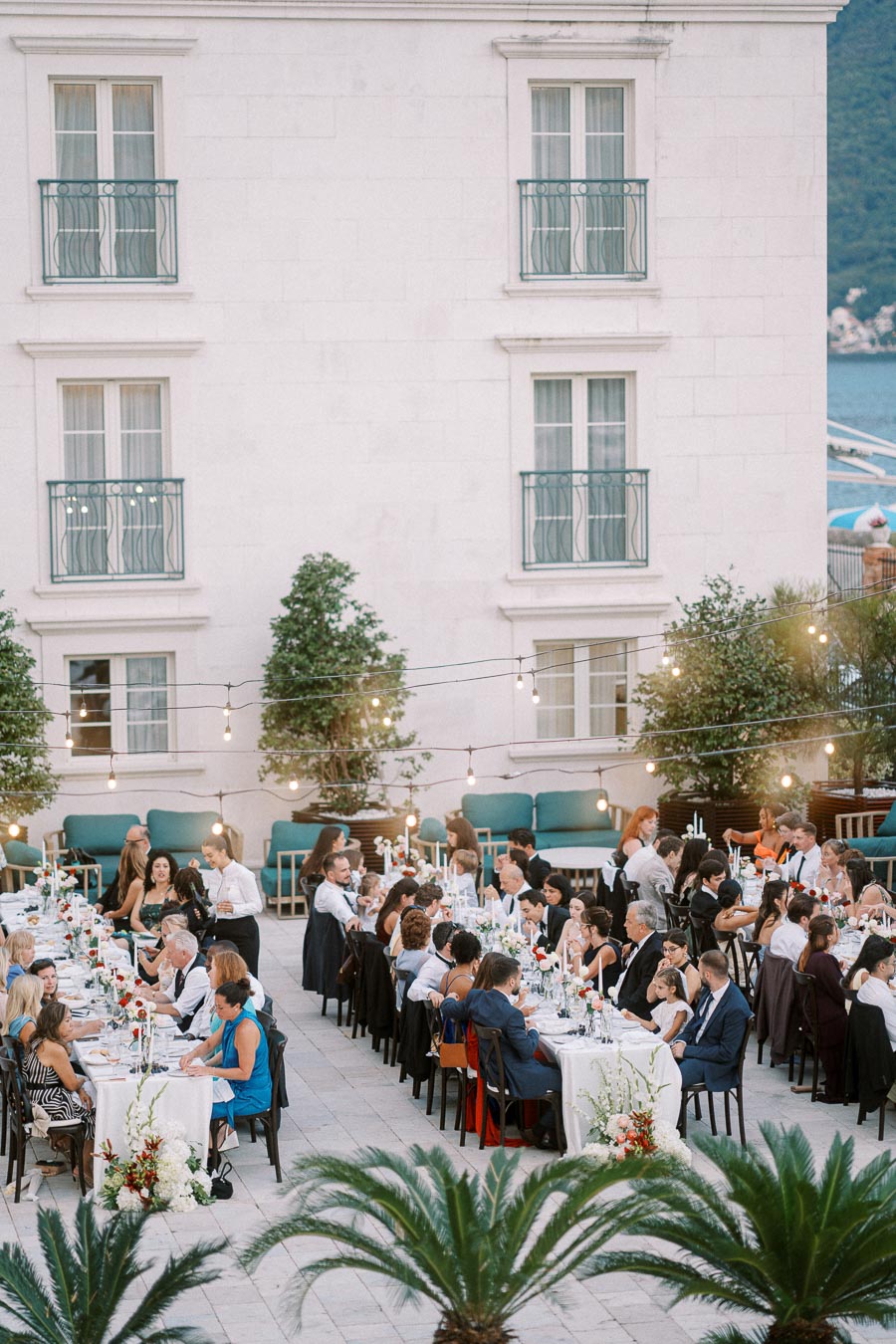 Elegant outdoor wedding reception with guests seated at long tables, surrounded by string lights and lush greenery in a charming courtyard setting.