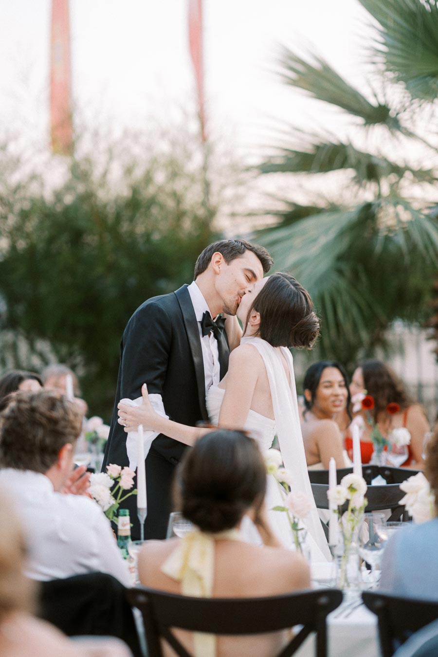 Newlywed couple sharing a romantic kiss at an elegant outdoor wedding reception, surrounded by guests and decorated tables.