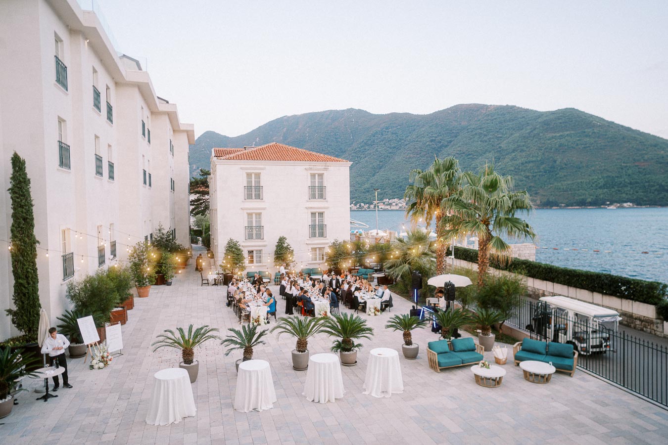 Elegant outdoor wedding reception by the sea with white table linens, palm trees, and mountain views; guests dining under string lights in a luxury coastal venue.