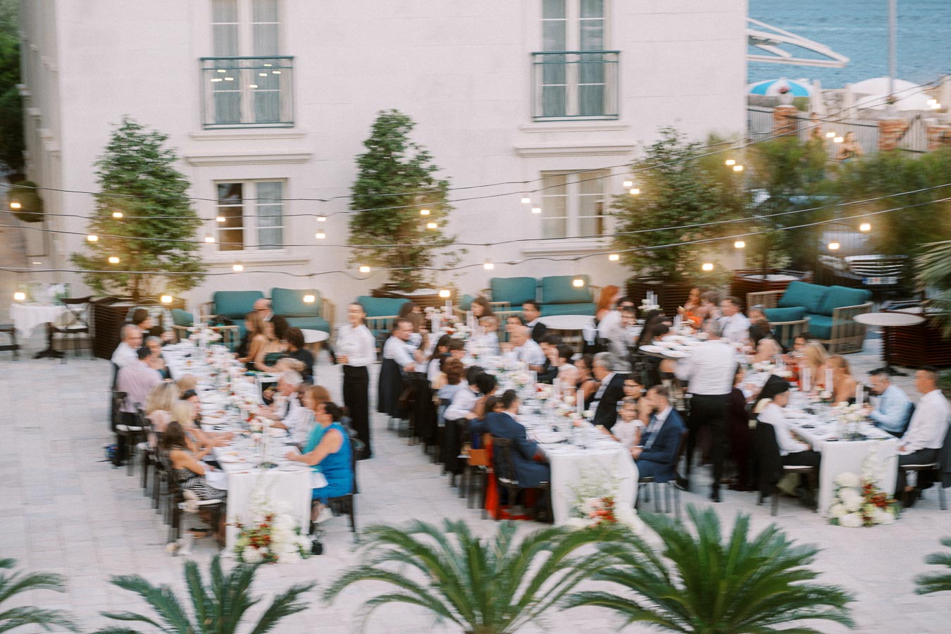 Outdoor wedding reception with guests seated at long tables decorated with floral arrangements, string lights overhead, and servers attending to the gathering in a courtyard setting.