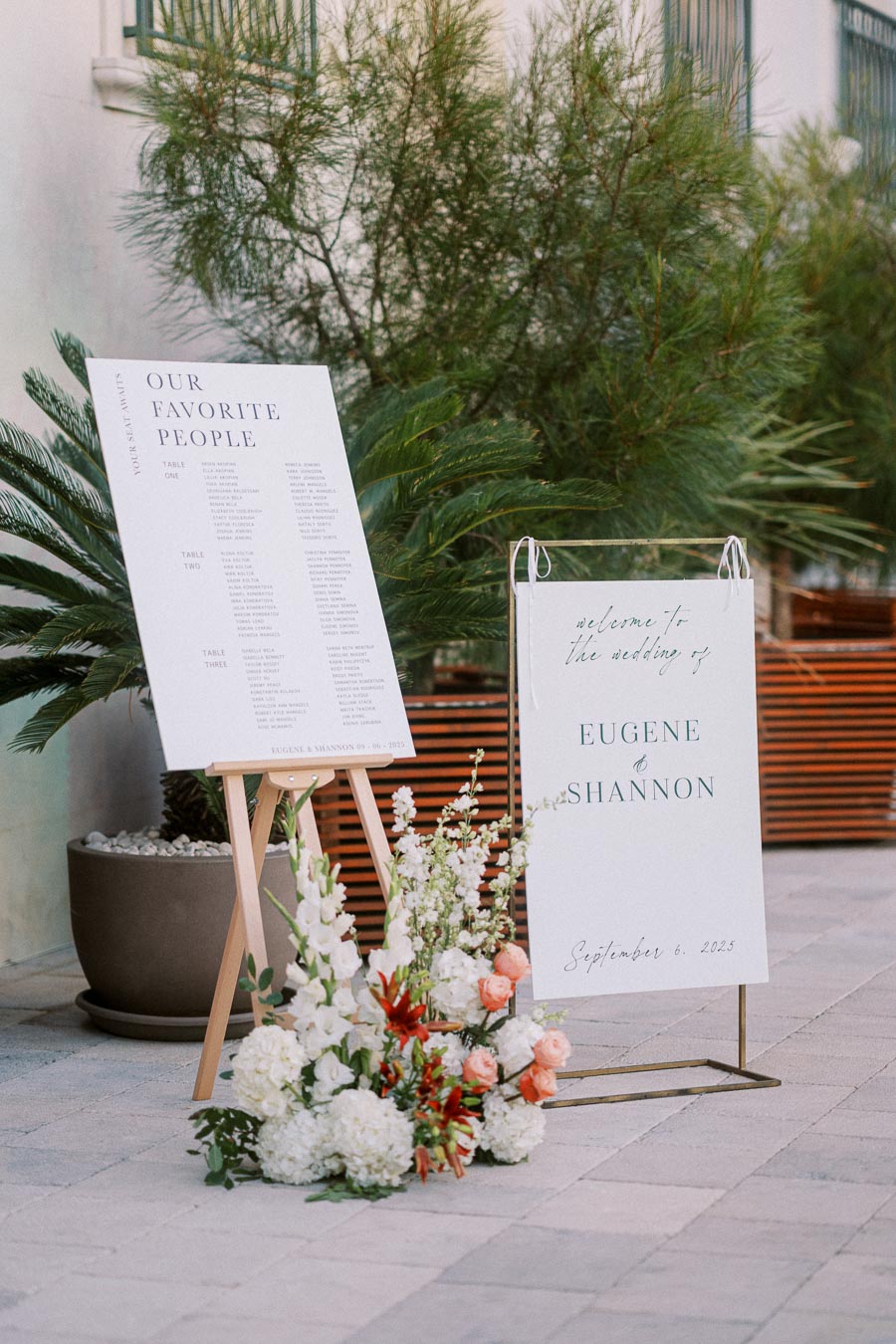 Elegant wedding welcome signs featuring guest seating arrangements and floral decor. The signs display information for Eugene and Shannon's wedding on September 6, 2025, set against a backdrop of lush greenery.
