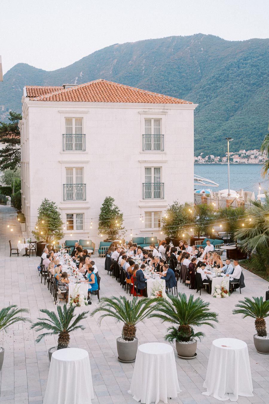 Elegant outdoor wedding reception with guests dining at long tables adorned with flowers and lights, set against a backdrop of a white building and scenic mountain views.