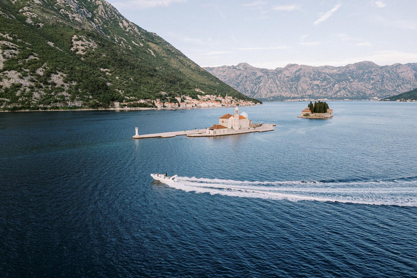 Aerial view of Our Lady of the Rocks island with a picturesque church in Montenegro's Bay of Kotor, surrounded by serene blue waters and mountainous landscape, with a speedboat passing by.
