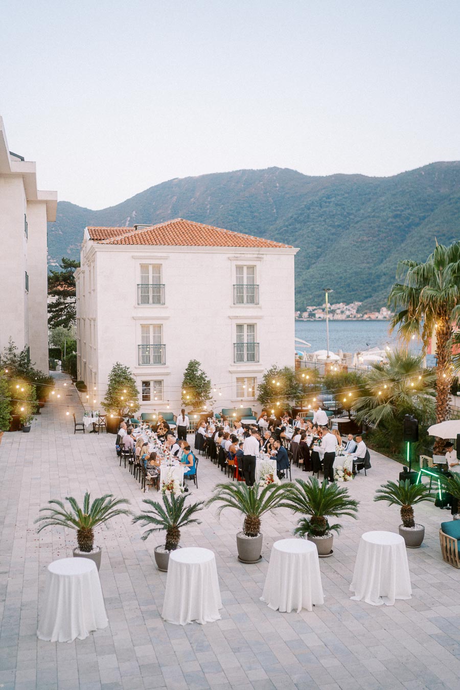Outdoor evening wedding reception in a courtyard with guests seated at long tables, surrounded by string lights and palm trees, against a backdrop of mountains and a historic building.