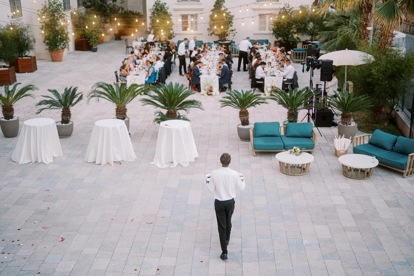 Outdoor wedding reception with elegant table settings, string lights above, and a waiter carrying drinks towards a seated crowd in a spacious courtyard adorned with palm trees.