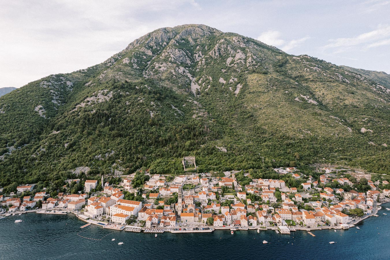 Aerial view of a coastal Mediterranean town with terracotta-roofed buildings nestled at the base of a lush green mountain, overlooking a calm blue sea.