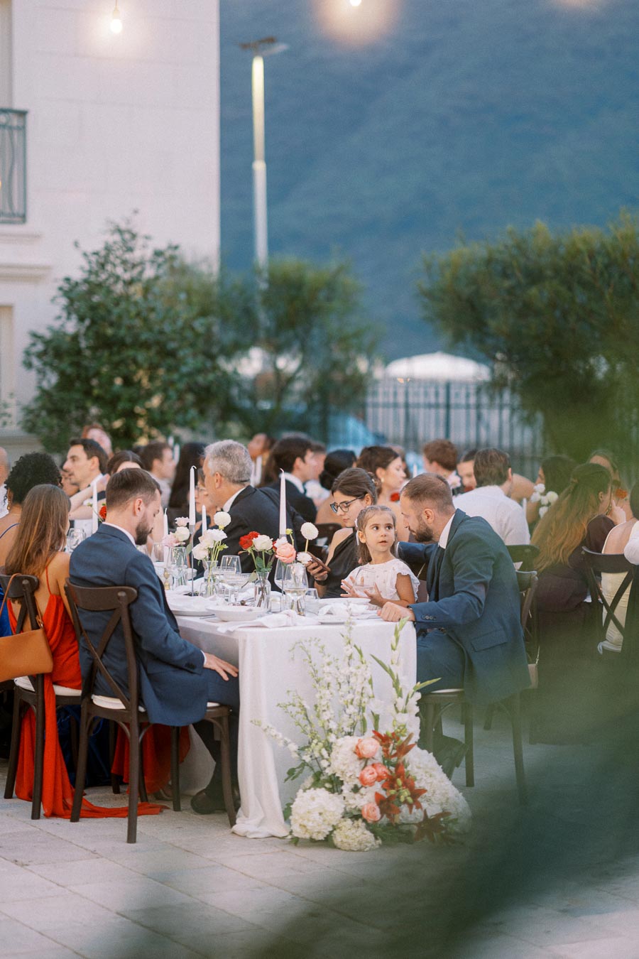 Outdoor wedding reception dinner with elegantly dressed guests seated at a long table adorned with white tablecloths, floral centerpieces, and candles, set against a scenic mountainous backdrop.