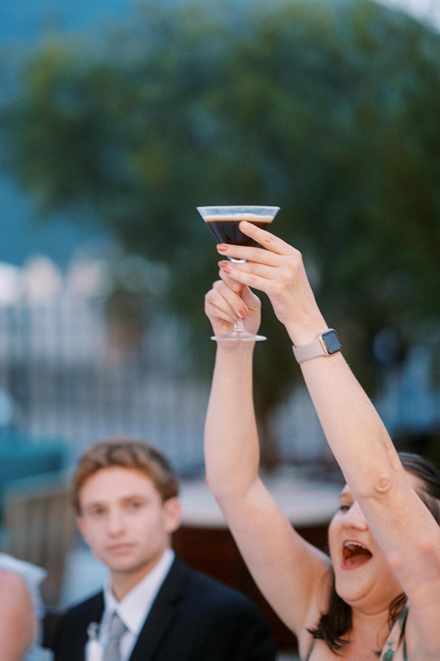 A person in a celebratory mood raising a dark cocktail in a martini glass, with an expression of joy, wearing an elegant outfit at an outdoor event.