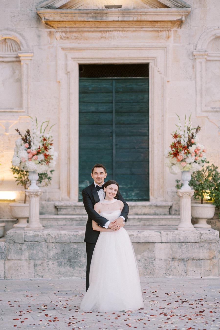 Elegant bride and groom pose together in front of historic building entrance, adorned with floral arrangements, on their wedding day.