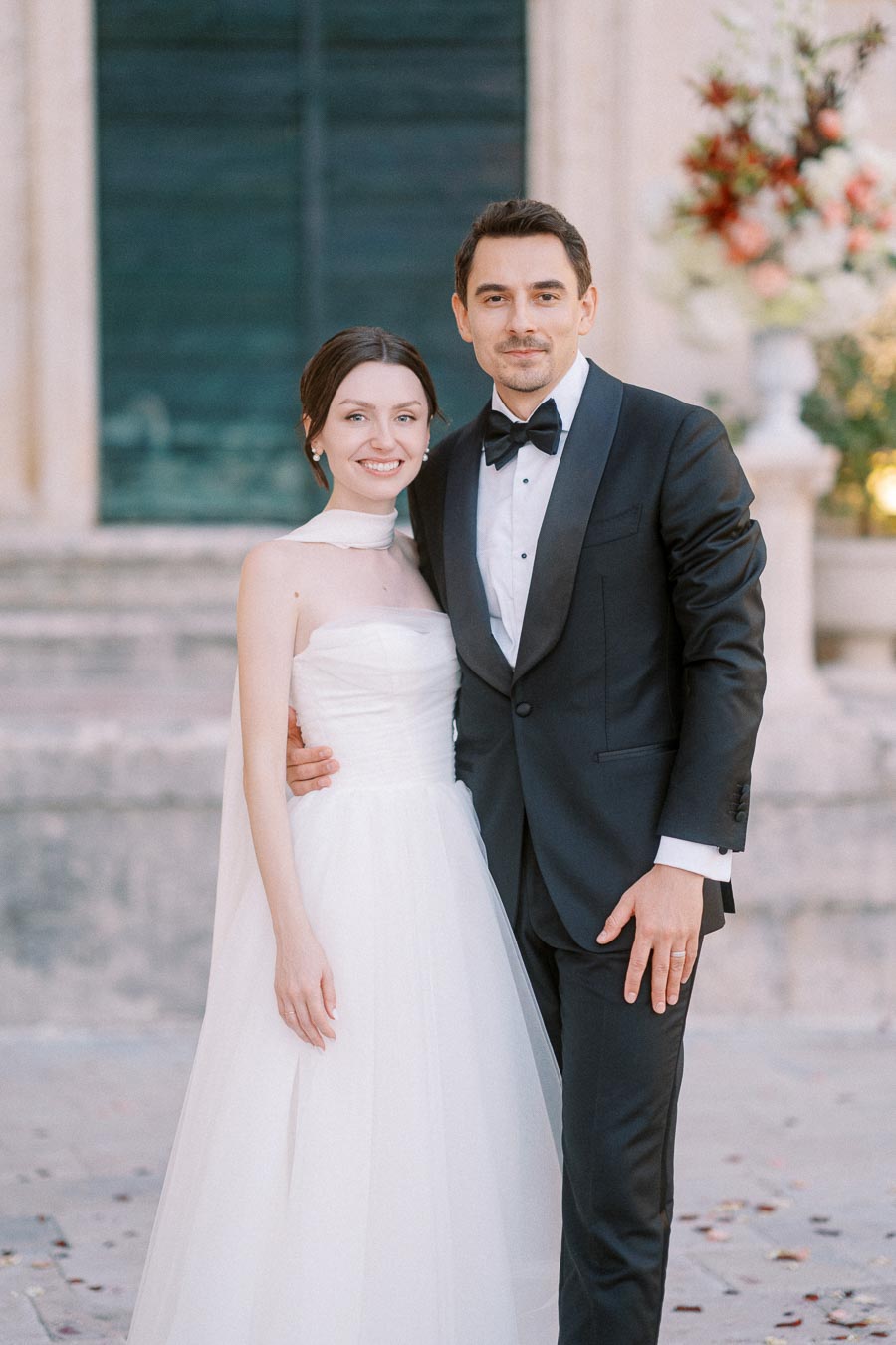 Elegant wedding couple posing in formal attire, with the bride in a strapless white gown and the groom in a classic black tuxedo, standing outdoors near stone steps and floral arrangements.