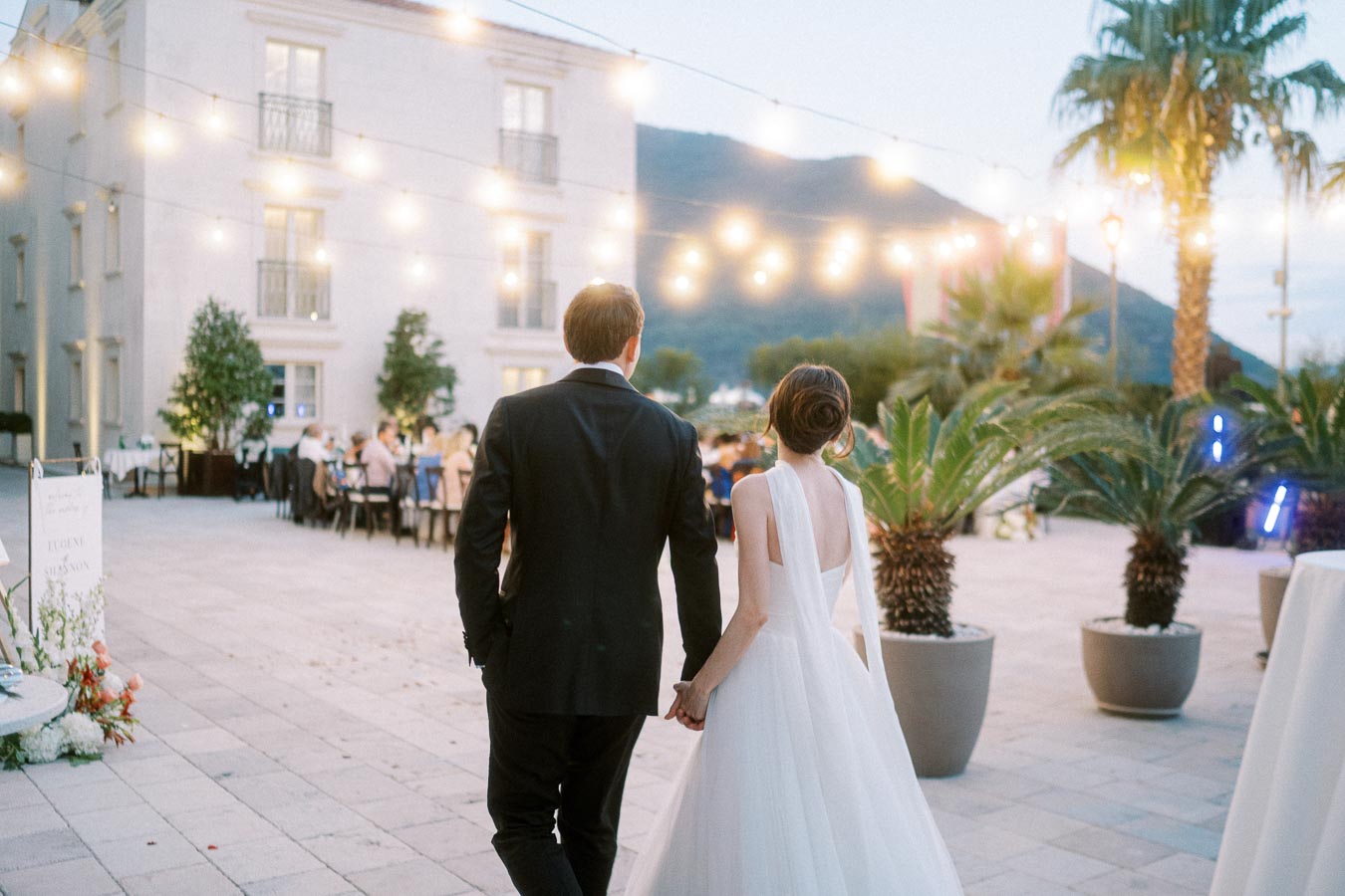 A couple in wedding attire walking hand in hand at an elegant outdoor evening reception, with string lights illuminating the courtyard and guests seated at tables in the background.