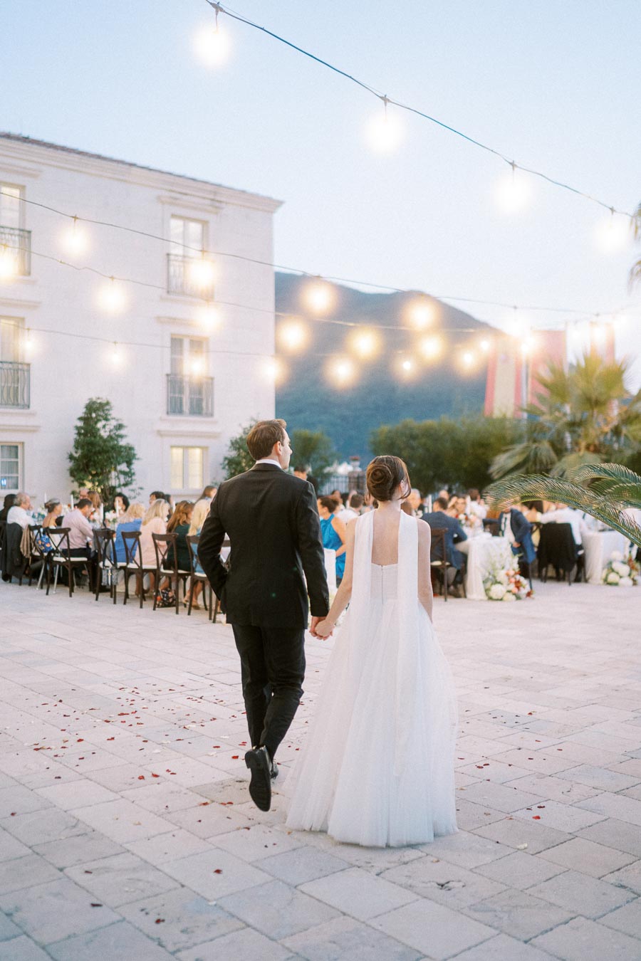 A bride and groom walk hand in hand towards a beautifully lit outdoor reception area, with guests seated at tables under string lights, creating a romantic wedding atmosphere.