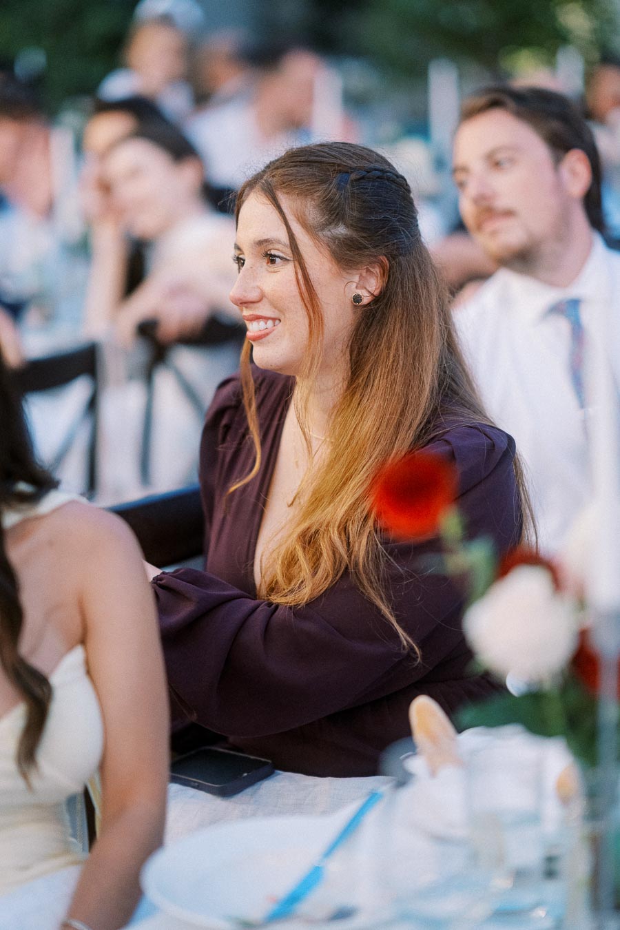 Smiling woman in a purple blouse at an outdoor event, sitting at a table with floral decorations and other attendees in the background.