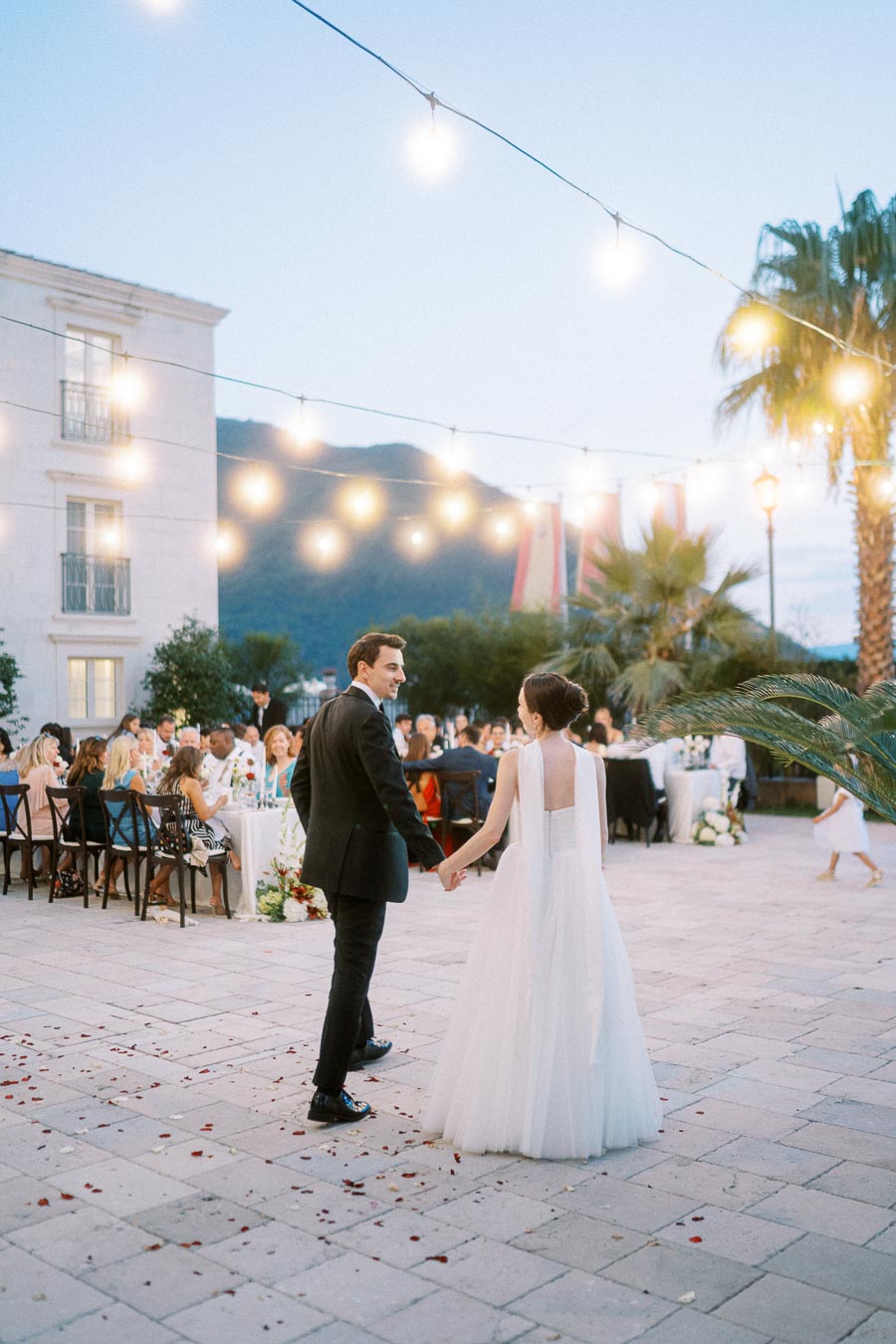 Elegant outdoor wedding reception with bride and groom under string lights, surrounded by guests at beautifully decorated tables.