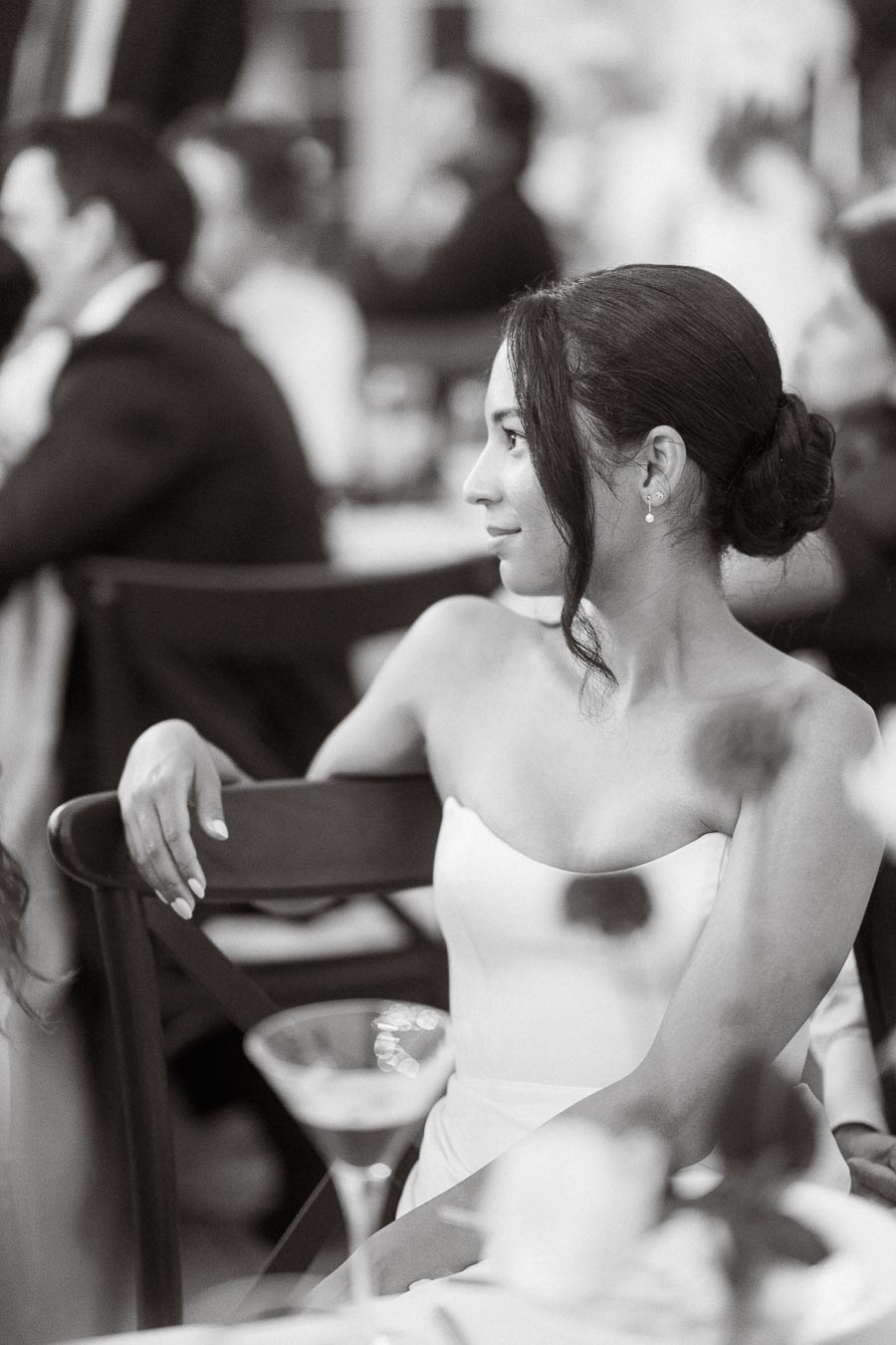 Elegant woman in strapless dress at formal event, seated with arm resting on chair, while holding a drink, in black and white.