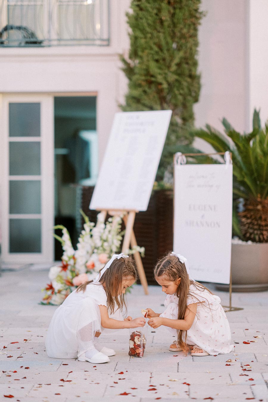 Two young flower girls in white dresses picking up petals outside at a wedding venue, with decorative signs and greenery in the background.
