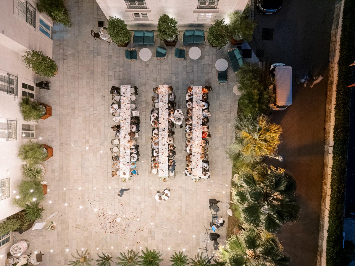 Aerial view of a beautifully decorated outdoor courtyard setting with long tables and guests at a nighttime event, surrounded by string lights and lush greenery.