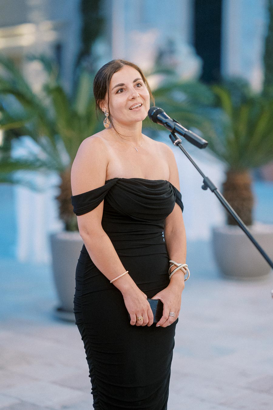 Elegant woman in a black dress speaking into a microphone at an outdoor event with palm plants in the background.