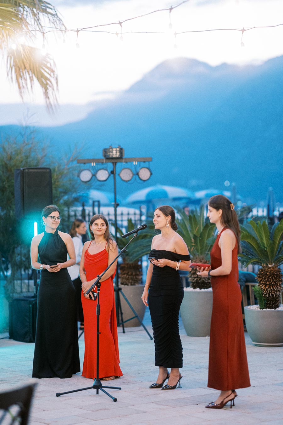 Four women in elegant evening gowns speaking at an outdoor event with mountains in the background, under hanging string lights.