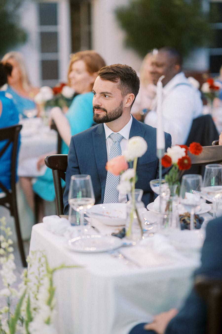 Man in a blue suit sitting at an elegant outdoor wedding reception, surrounded by guests and floral table settings.