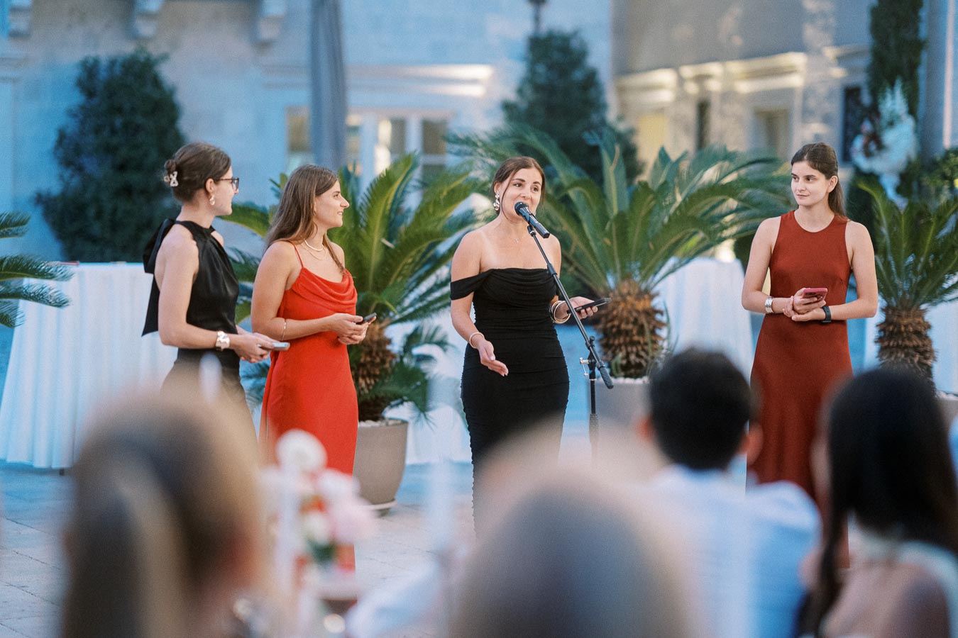 Four women elegantly dressed in evening gowns speaking and holding smartphones at an outdoor event, surrounded by potted plants and facing an audience.