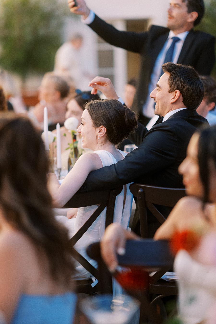 A joyful wedding reception with a couple smiling and seated, surrounded by guests in an outdoor setting.