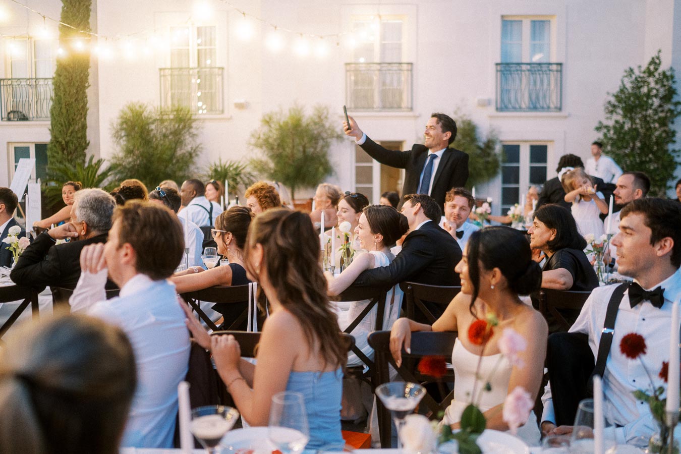 Elegant outdoor wedding reception with guests sitting at tables under string lights, as a man in a suit takes a photo with a smartphone.