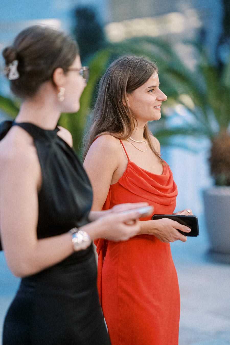 Two women at an elegant event, one wearing a red dress and holding a phone, the other in a black dress, both appearing engaged and smiling outdoors.