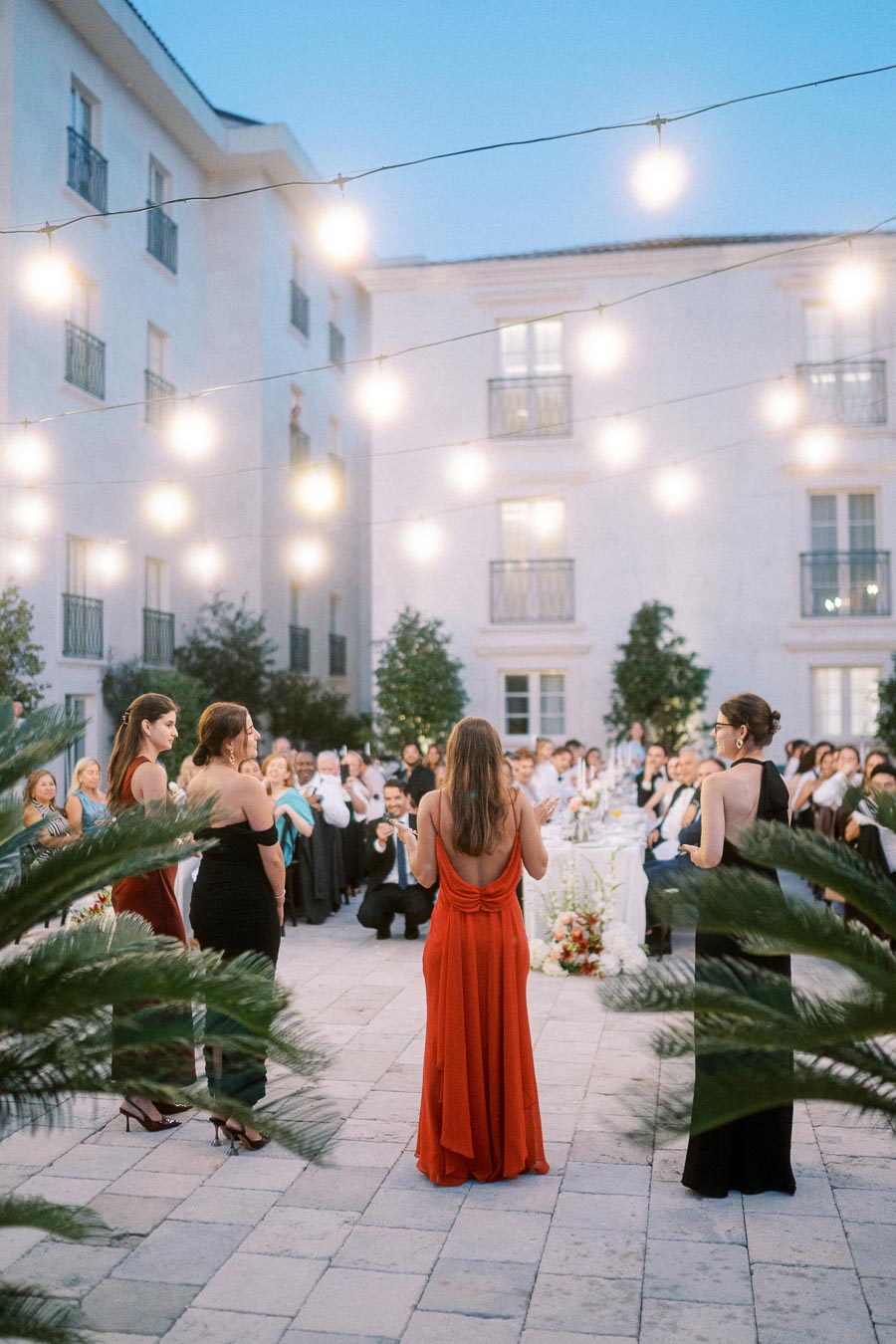 Elegant outdoor wedding reception with women in evening gowns speaking to guests under string lights, surrounded by lush greenery and a beautifully set dinner table.