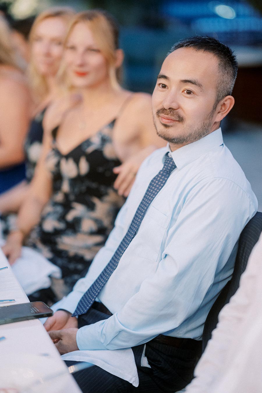 A person is seated at a formal event, wearing a light blue shirt and a patterned tie, with others sitting in the background.