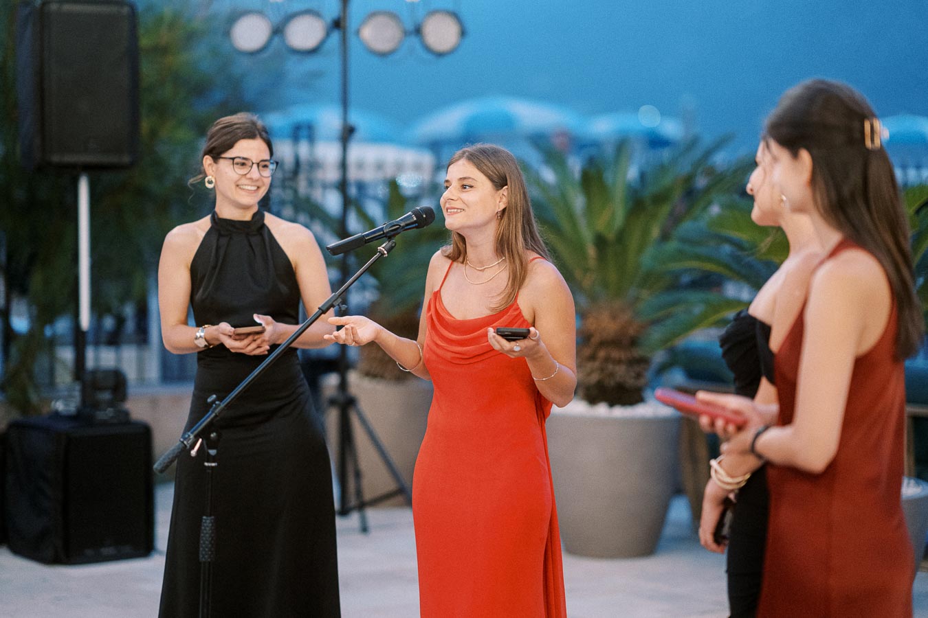 Women in elegant evening dresses speaking into a microphone at an outdoor event, surrounded by plants and soft background lighting.