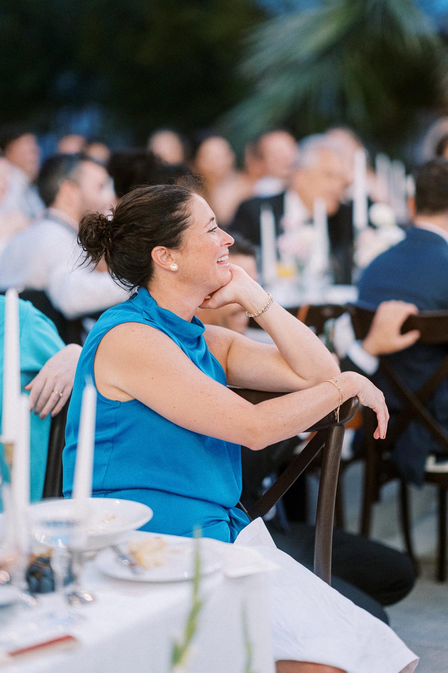A woman in a blue dress smiling at a formal event, seated at a table adorned with white candles and dinnerware, surrounded by guests.