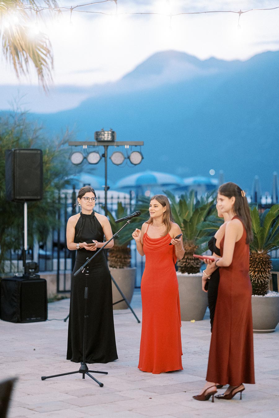 Three women in elegant evening gowns presenting with microphones at an outdoor event against a scenic mountain backdrop at dusk.