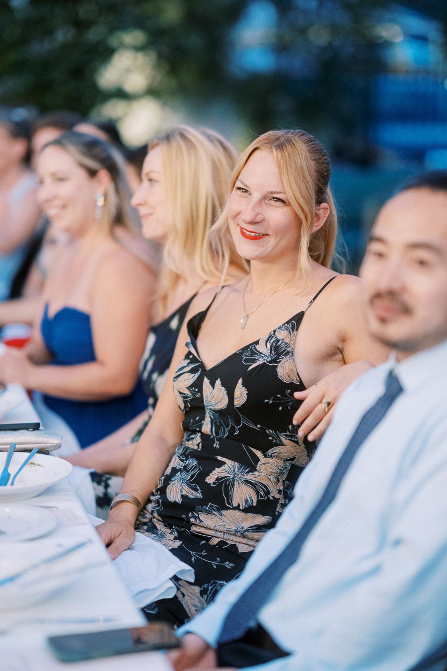 A group of people smiling and seated at a formal outdoor event, with a woman in a floral dress in focus.