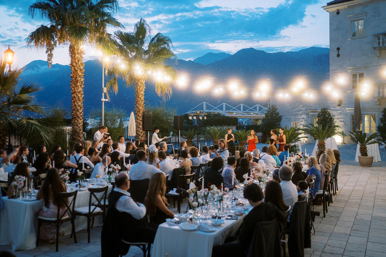 Outdoor evening wedding reception with guests dining under string lights, palm trees, and a scenic mountain backdrop.