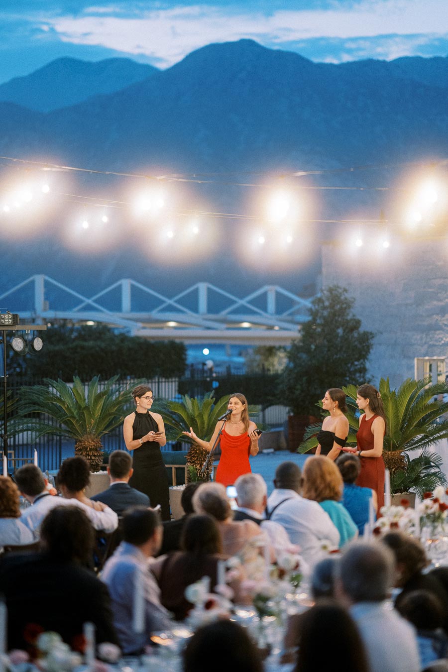 Outdoor evening wedding reception with four women speaking to guests under string lights, with a mountainous backdrop.