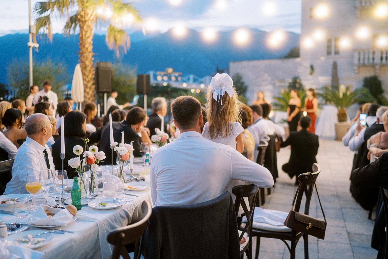 Outdoor wedding reception at sunset with elegantly dressed guests seated at decorated tables, twinkling string lights above, and scenic mountain view in the background.