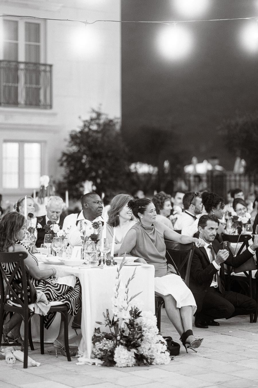 Black and white photo of elegantly dressed guests seated at an outdoor wedding reception, attentively listening to speeches, with floral arrangements and string lights creating a sophisticated atmosphere.