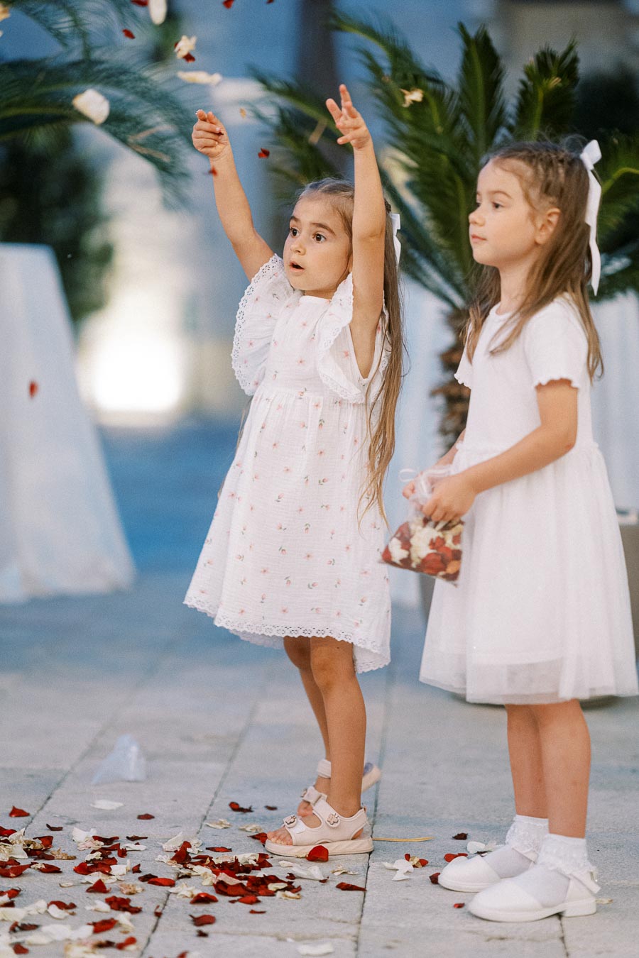 Two young girls in white dresses playfully scatter flower petals at an outdoor event, surrounded by greenery and a soft, festive ambiance.