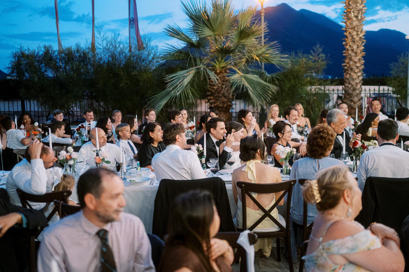 Outdoor evening event with elegantly dressed guests seated at round tables, surrounded by palm trees, under a twilight sky.
