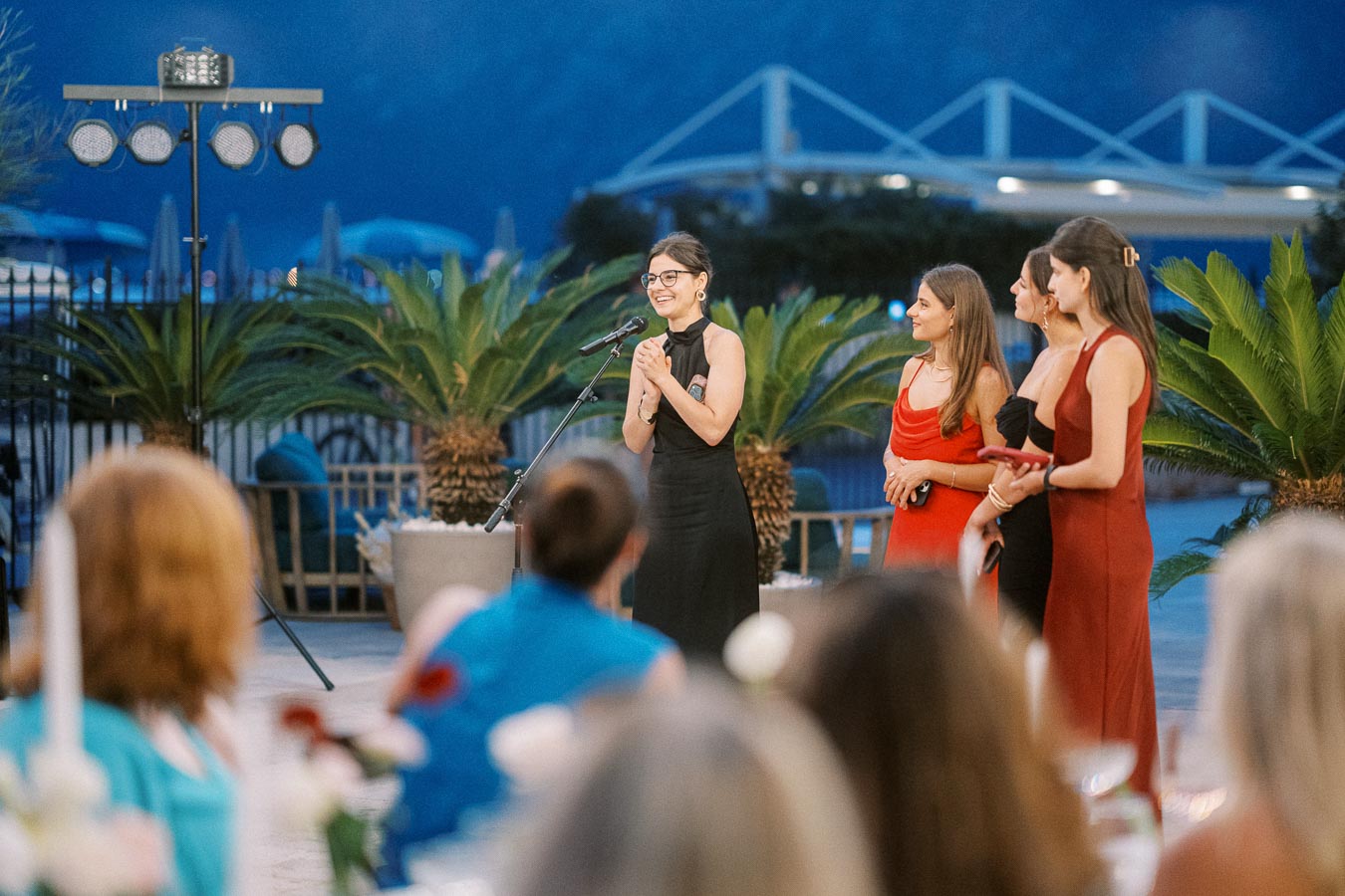 Four women elegantly dressed in evening gowns stand together on an outdoor stage, illuminated by event lighting, with a tropical backdrop under the night sky.