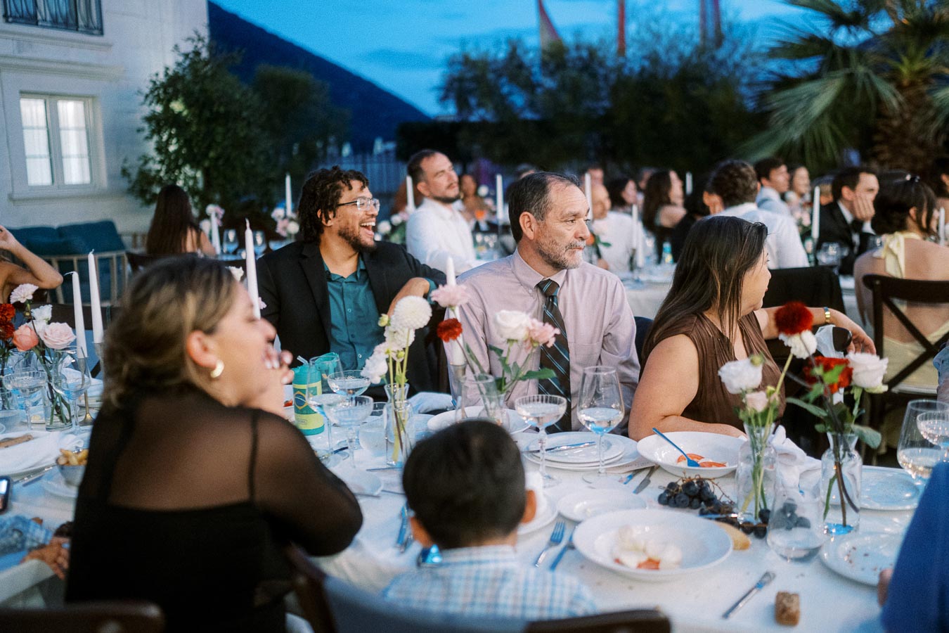 Group of people enjoying a lively outdoor dinner gathering with floral arrangements and dining setup.