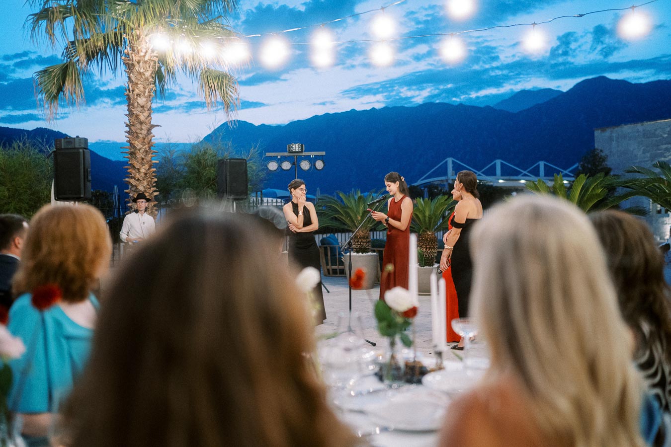 Outdoor evening event with people gathered around a table, listening to a speech. Three individuals are at the front under string lights, with a scenic backdrop of mountains and palm trees, creating a festive and elegant atmosphere.