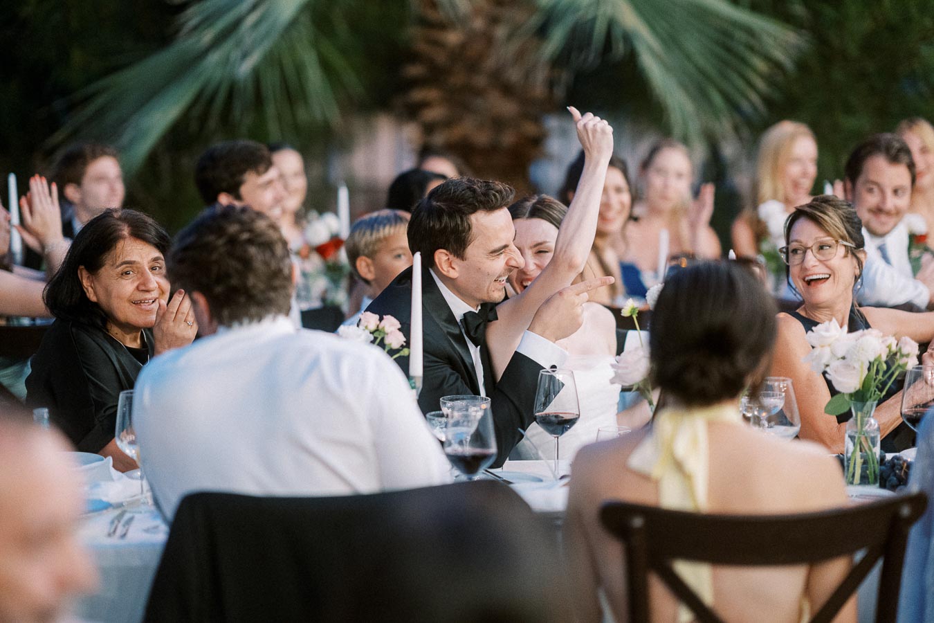 Guests enjoying a joyful moment during an outdoor wedding reception, seated at a table with elegant decor, including flowers and wine glasses, with palm trees visible in the background.